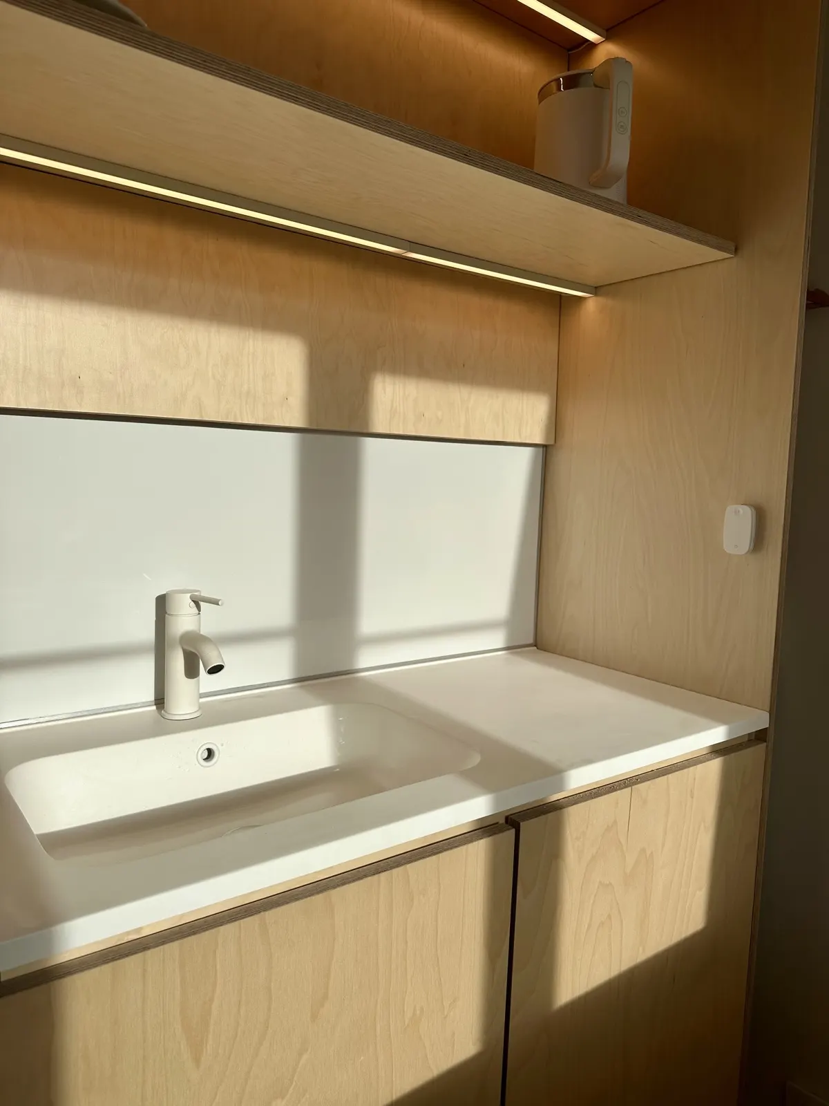Vasque et plan de toilette blancs sous un bandeau lumineux, menuiserie bouleau
